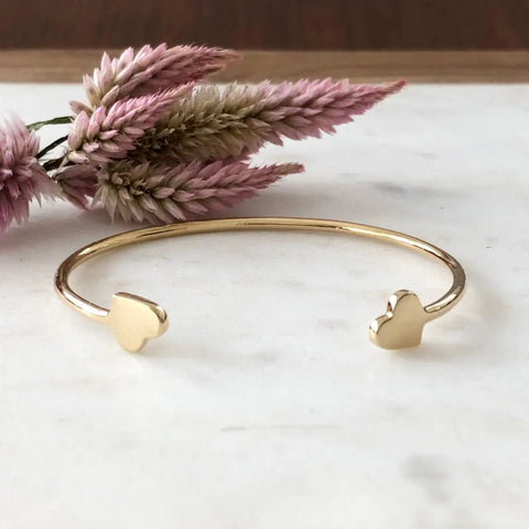 The GOLD VALEN BRACELET by Sarah Cecelia, a gold cuff with heart-shaped ends, is displayed on white marble beside pink and beige dried flowers—a minimalist piece thats a perfect gift for her.