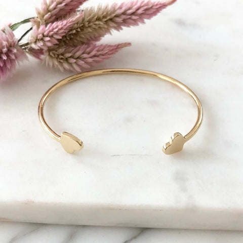 The GOLD VALEN BRACELET by Sarah Cecelia is a minimalist gold cuff with two small heart-shaped ends, shown on white marble with soft pink dried flowers—the perfect gift for her.