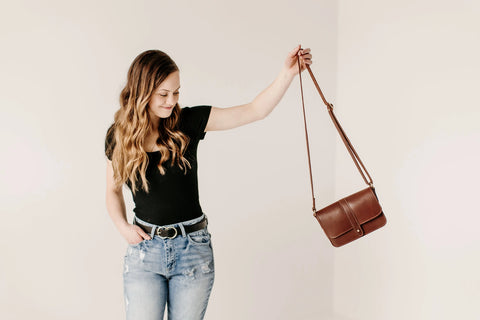 A woman with long wavy hair in a minimalist black t-shirt and blue jeans stands with one hand in her pocket, smiling slightly as she holds up the Urban Southern Lancaster 5-Pocket Crossbody against a plain background.
