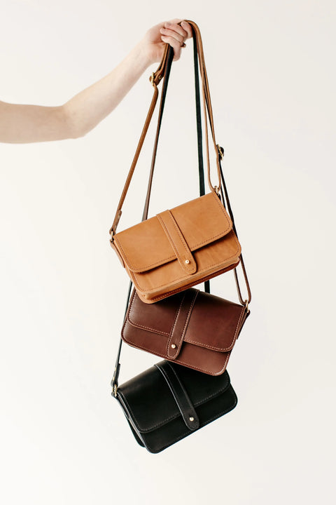 A hand displays three Urban Southern Lancaster 5-Pocket Crossbody bags in tan, brown, and black full-grain leather with front flaps and button closures, bringing minimalist style to a plain light background.
