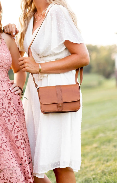 Two women stand on grass in a blurred green setting; one, dressed in a minimalist white short-sleeve dress, carries an Urban Southern Lancaster 5-Pocket Crossbody bag, while the other, partly visible, wears a pink lace dress.