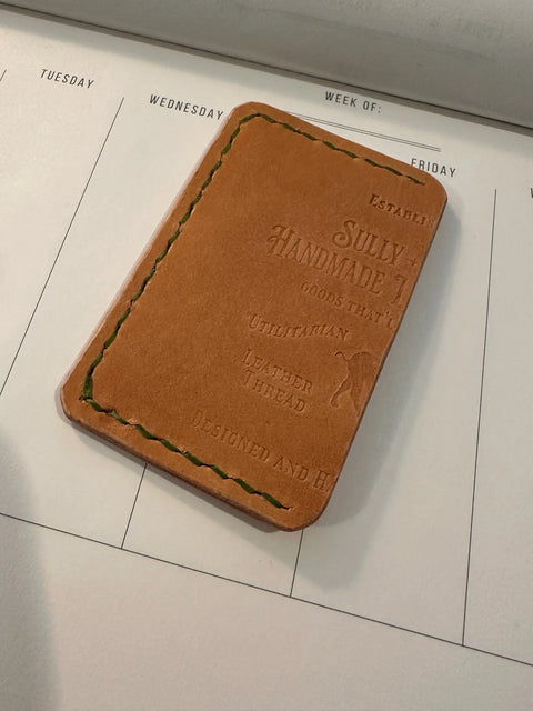 A tan Cash Stash Sleeve - Small by Sully + Proper Supply, crafted from upcycled leather with green stitching, sits on a weekly calendar. Handmade in the USA, this wallet is embossed with decorative text and patterns.