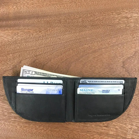 A Nantucket Front Pocket Wallet by Rogue Industries lies open on a wooden surface, showing several credit cards, a Maine driver’s license, and folded US dollar bills inside.