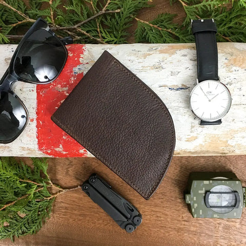 A Rogue Industries Nantucket Front Pocket Wallet, black sunglasses, a silver wristwatch with a black strap, a black multitool, and a camouflage-patterned pouch are arranged on a wooden surface with green foliage.