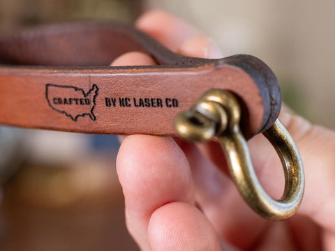A hand holds the Minimalist Leather Key Fob – Horseshoe Shackle Design, made in the USA by KC Laser Co. The leather strap features an outline of the US map and CRAFTED BY KC LASER CO, highlighting its American-made quality.