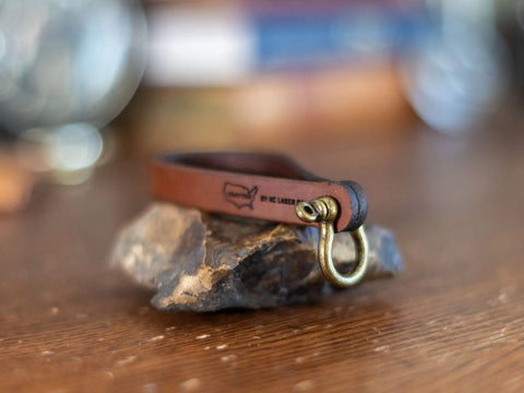 The Minimalist Leather Key Fob by KC Laser Co features a solid brass horseshoe shackle and USA logo, displayed on stone over wood with a blurred background. Made in the USA.