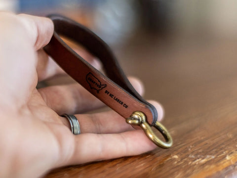 A hand holds the Minimalist Leather Key Fob – Horseshoe Shackle Design by KC Laser Co, crafted in the USA. Featuring durable leather, a solid brass horseshoe shackle, and a U.S. stamped design, it embodies classic style and quality craftsmanship.