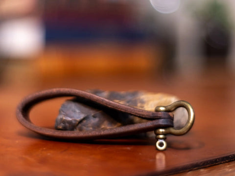 The Minimalist Leather Key Fob – Horseshoe Shackle Design by KC Laser Co sits on a polished wood surface with a blurred background and a partially visible stone behind it. Made in the USA.