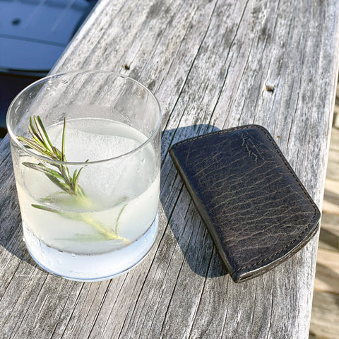A clear cocktail with ice and rosemary rests on weathered wood beside The Minimalist Wallet by Rogue Industries, illuminated by bright sunlight.