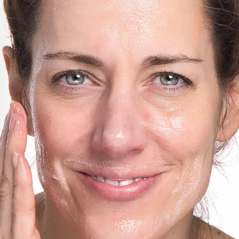 A woman smiles gently while applying Open Formula Oil Cleanser to her face, her skin glistening as she uses her fingers to spread the product, enjoying a deep cleanse that leaves no oily residue.