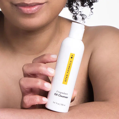 A person with curly hair holds a white Open Formula Oil Cleanser bottle close to their chest, displaying this effective pore-cleansing oil and makeup remover against a plain white background.