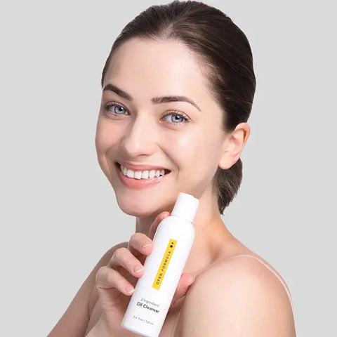A smiling woman with light eyes and brown hair holds a white Open Formula Oil Cleanser bottle with yellow label, posing against a plain light gray background.