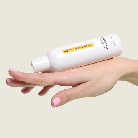 A hand with light pink nails balances a white Open Formula Oil Cleanser bottle on the back of the fingers against a neutral background; this oil cleanser removes makeup and deeply cleanses pores.