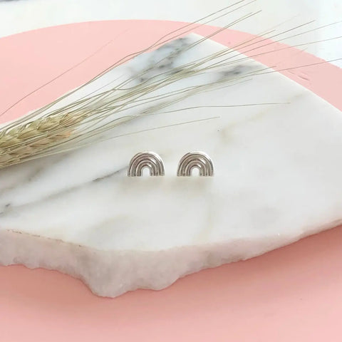 The SILVER ARCO STUDS by Sarah Cecelia, minimalist sterling silver rainbow earrings, rest on a white marble slab with a pink background and a dried wheat stem nearby.