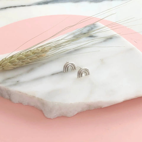 The SILVER ARCO STUDS by Sarah Cecelia, two small sterling silver rainbow earrings, are shown on a white marble slab beside dried wheat and set against a pastel pink background, highlighting their modern geometric design.