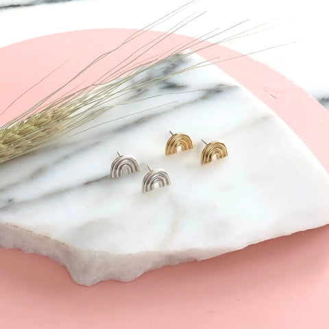 The Sarah Cecelia SILVER ARCO STUDS, a pair of geometric rainbow-shaped sterling silver earrings, are showcased on white marble with a pink background and dried wheat—ideal for minimalist jewelry lovers.