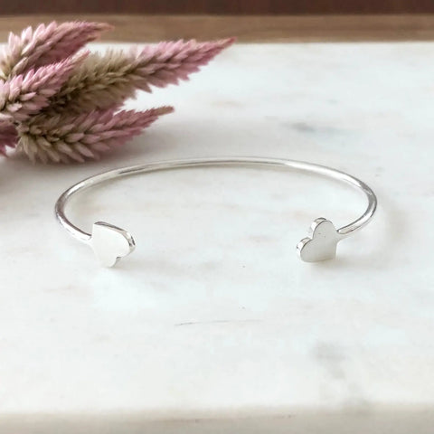 The SARAH CECELIA SILVER VALEN BRACELET, featuring heart-shaped ends, is displayed on white marble with soft pink dried flowers in the background, emphasizing its minimalist charm.