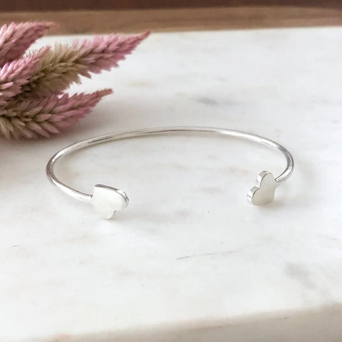 The SILVER VALEN BRACELET by Sarah Cecelia, a handmade silver piece with heart-shaped ends, lies on white marble with pink dried flowers in the background, showcasing its minimalist charm.
