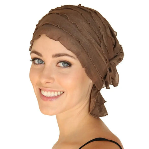 A smiling woman with light skin and blue eyes wears the Chemo Beanies® Sara textured brown chemo beanie and a black strap top, facing sideways with her head turned slightly toward the camera against a white background.