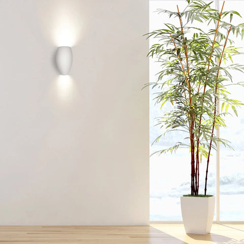 A tall green potted plant sits on a light wood floor beside a white wall illuminated by the handcrafted Modern USA-Made Aruba Sconce from A19 Artisan Lighting, casting light up and down. Sunlight streams in from a window to the right.