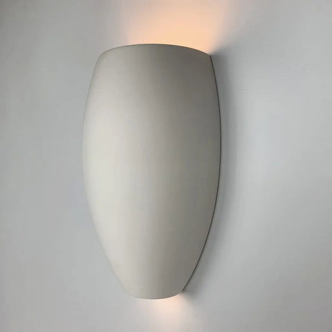 The Aruba Sconce from A19 Artisan Lighting is a handcrafted, modern wall light made in California, featuring an oval shape in white that casts gentle up and down lighting on a light grey wall.