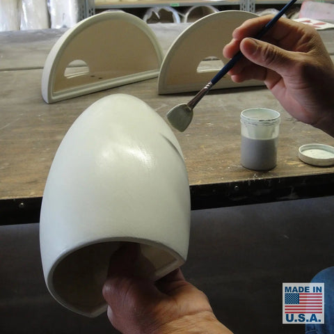 A person paints a white ceramic Modern USA-Made Wall Lighting – Aruba Sconce, handcrafted in California by A19 Artisan Lighting. Modern wall sconces and paint are on the table, with a Made in U.S.A. label visible nearby.