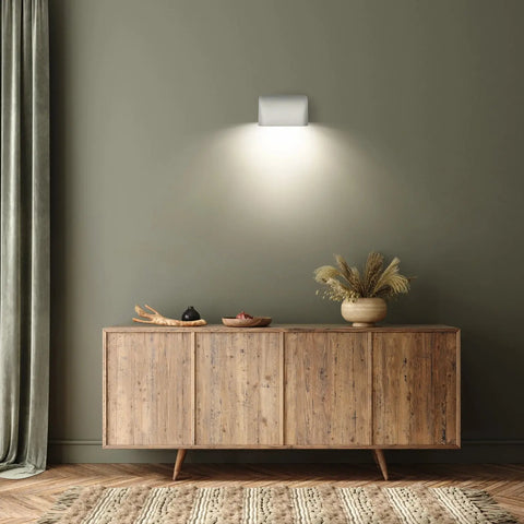 A wooden sideboard with decorative bowls and a vase of dried grass stands against a green wall, illuminated by the Balboa Downlight Sconce from A19 Artisan Lighting, handcrafted in California. Draped curtains and a textured rug complete the minimalist room decor.
