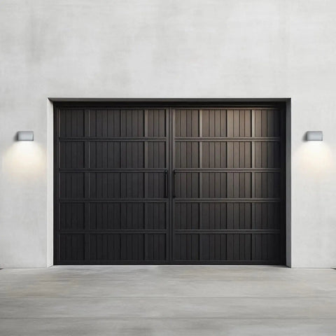 A modern black double garage door with vertical panels is set in a smooth light gray concrete wall. On either side, A19 Artisan Lightings USA-made Balboa Downlight Sconce casts soft, symmetrical glows. Handcrafted in California.