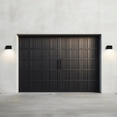 A modern, dark gray double garage door with vertical and horizontal panels is centered on a plain, light wall. Flanking it are two A19 Artisan Lighting Balboa Downlight Sconces with LED Smart Bulbs. The driveway is concrete.