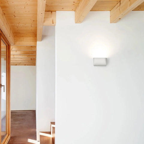 A modern interior with white walls, exposed wooden beams, a wood-framed glass door, and the Balboa Downlight Sconce by A19 Artisan Lighting handcrafted in California. Wooden stairs are partially visible in the lower right corner.