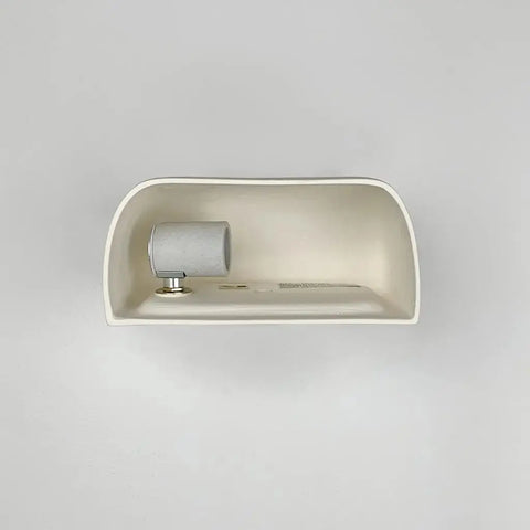 A white, minimalist wall-mounted shelf holds the Modern USA-Made Balboa Downlight Sconce by A19 Artisan Lighting—handcrafted in California—plus a metallic button, all set against a plain white background.