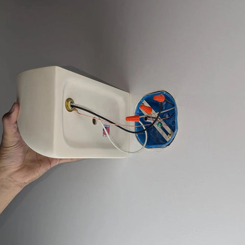 A hand holds the Balboa Downlight Sconce by A19 Artisan Lighting, a modern wall-mounted fixture handcrafted in California, next to an electrical box with exposed wires secured by orange wire nuts on a light-colored wall.