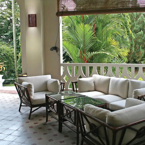 Outdoor patio with cushioned rattan chairs, a glass coffee table, and lush greenery. Tiled floor and partial white railing. Handcrafted in California by A19 Artisan Lighting, the USA-made Bamboo Sconce adds energy-efficient LED light for a relaxing vibe.