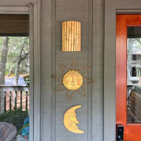 A gray wall on a porch displays the Modern USA-Made Wall Lighting – Bamboo Sconce by A19 Artisan Lighting above golden sun and moon metal decor, next to a wooden-framed glass door.