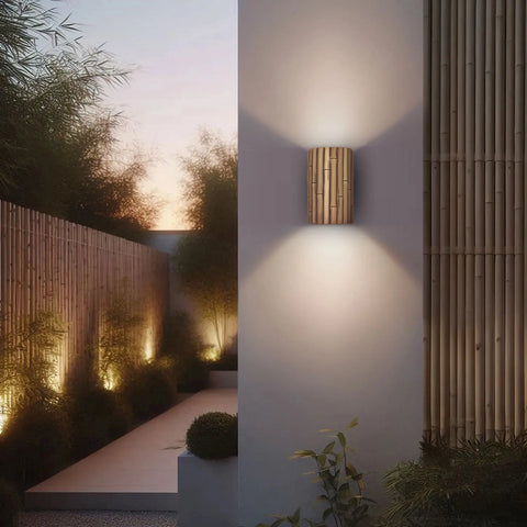 At dusk, a modern outdoor space with bamboo fencing and bushes is lit by A19 Artisan Lightings Modern USA-Made Bamboo Sconce, handcrafted in California, casting a warm glow on the white wall while path lights highlight the garden.