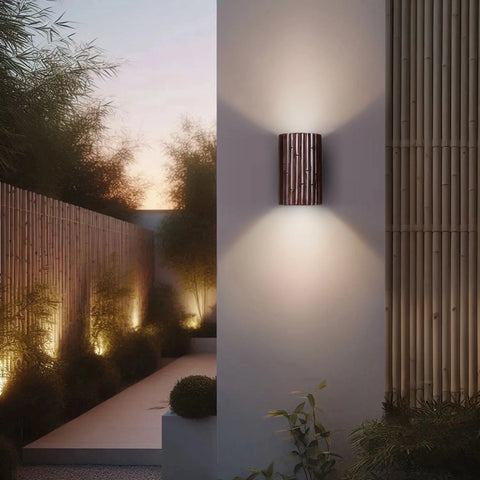 The Modern USA-Made Wall Lighting – Bamboo Sconce by A19 Artisan Lighting, handcrafted in California, features a cylindrical slatted design and casts a warm glow against a white garden wall with bamboo fencing and LED pathway lights at dusk.