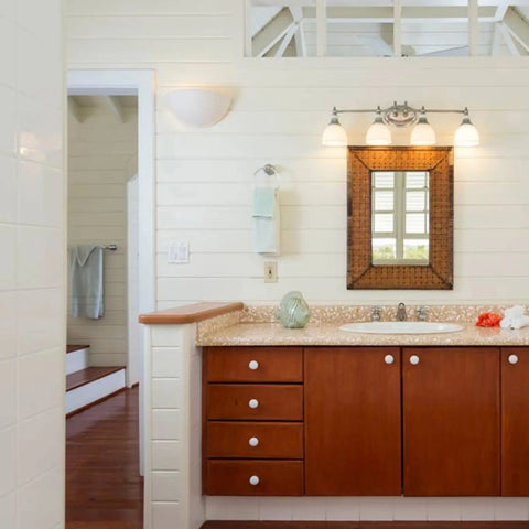 A bathroom features a wooden vanity with a granite countertop, a rectangular mirror topped by Capri Sconce wall lighting from A19 Artisan Lighting, a sink, white paneled walls, and a hand towel beside the mirror. Another room is visible in the background.