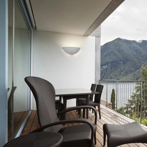 A modern balcony with wicker chairs and a table on wood flooring overlooks mountains and a lake. A19 Artisan Lighting’s Capri Sconce, handcrafted in California, glows on the white wall beside large glass doors.