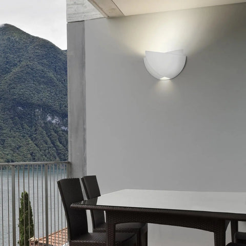 A modern patio features a dark wicker table and chairs, a gray wall with the Modern USA-Made Ceylon Sconce by A19 Artisan Lighting, handcrafted in California, and scenic green mountains with a lake in the background.