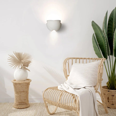 A cozy corner features a wicker chair, white textured pillow and blanket, plus a wicker side table and decorative vase. A large green plant accents the wall, illuminated by the Ceylon Sconce from A19 Artisan Lighting, handcrafted in California.