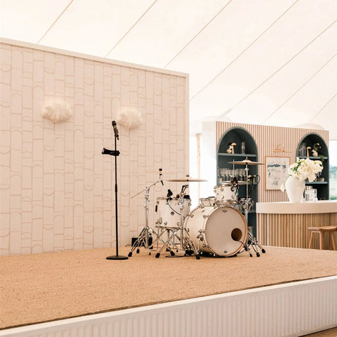 A white drum set and microphone stand sit on a beige carpeted stage, illuminated by Modern USA-Made Wall Lighting – Fiji Sconce from A19 Artisan Lighting, in a bright venue with a bar and floral arrangements in the background.