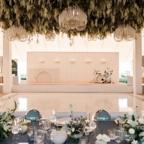 A chic event space with a white stage, drum set, greenery, chandeliers, glossy dance floor, and blue-table settings—glowing with the Modern USA-Made Fiji Sconce handmade by A19 Artisan Lighting in California.