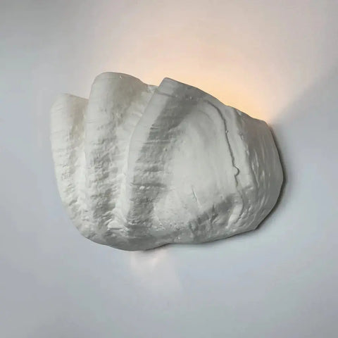 The Modern USA-Made Wall Lighting – Fiji Sconce by A19 Artisan Lighting is handcrafted in California, shaped like a seashell, and emits a soft, warm glow against a light-colored wall.