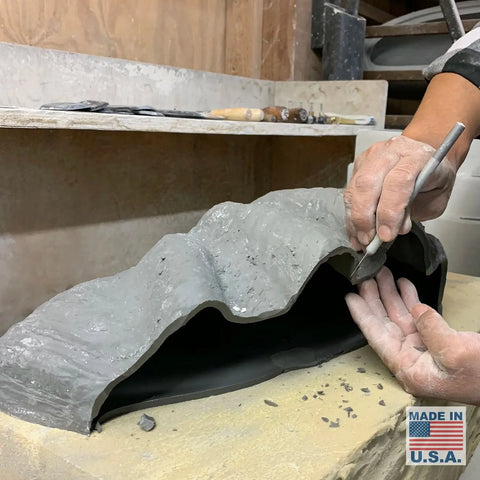 An artisan sculpts a piece by hand on a workbench, surrounded by carving tools and dust. A “Modern USA-Made Wall Lighting – Fiji Sconce” from A19 Artisan Lighting, handcrafted in California, lights the scene with energy-efficient LED.
