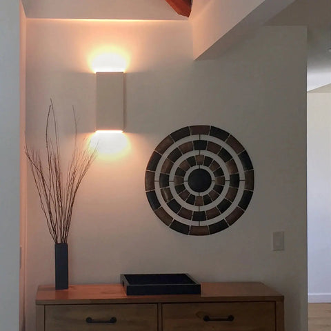 A19 Artisan Lighting’s USA-made Flores Sconce, handcrafted in California, casts light above a circular wall art. Below, a wooden dresser holds a dark vase with tall branches and a tray, creating a minimalist contemporary look.