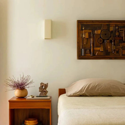 A minimalist bedroom features a single bed with beige bedding, a wooden headboard, and the Modern USA-Made Wall Lighting – Flores Sconce by A19 Artisan Lighting, handcrafted in California, softly illuminating a wooden nightstand with decor.