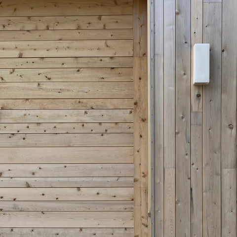 A19 Artisan Lighting’s Modern USA-Made Flores Sconce, handcrafted in California, is mounted on vertical light wood planks beside horizontal planks. The wall shows natural knots and grain; the sconce uses LED smart bulbs.