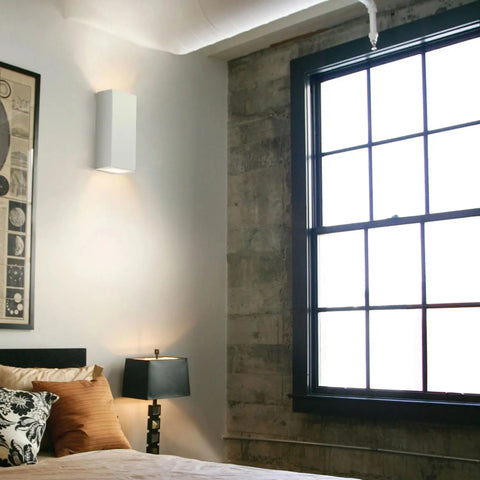 A modern bedroom corner with a bed, decorative pillows, abstract wall art, and a large black-framed window. The space features white and concrete walls plus the Flores Sconce by A19 Artisan Lighting, handcrafted in California.