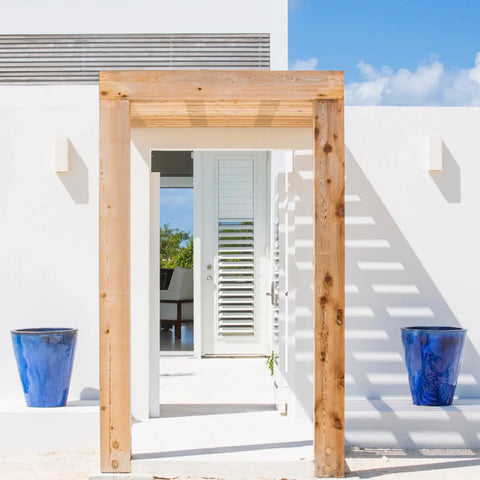 A modern white building with a wooden archway, blue ceramic pots, and a white door with plantation shutters is illuminated by Modern USA-Made Wall Lighting – Flores Sconce, handcrafted in California by A19 Artisan Lighting.