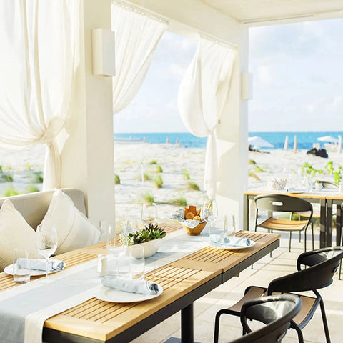 A bright seaside restaurant with wooden tables, sheer white curtains, and cushioned seating overlooks the ocean—illuminated by Modern USA-Made Flores Sconces, handcrafted in California by A19 Artisan Lighting, for a custom ambiance.